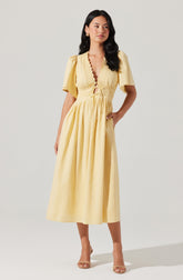 A woman is wearing a long, yellow dress made of a lightweight, breezy fabric. The dress features short puffed sleeves, a deep V-neckline with a scalloped edge, and a drawstring waist that creates a gathered effect. The skirt is full and flows down to her ankles. She is also wearing nude high-heeled sandals.